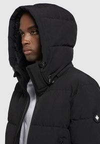 Schwarze Steppjacke mit einem genähten Design, großer Kapuze und Kordelzug. Enthält ein Logopatch am linken Arm und einen Reißverschluss vorne.