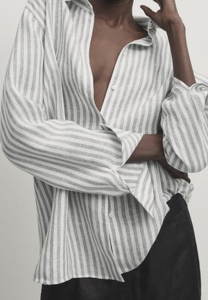 Person wearing loose white and gray vertically striped button-up shirt with open collar and black pants, arm crossed and hand near face.