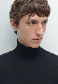 Svart polotröja av slätt tyg, tajt passform runt halsen, minimal design; modellen har ljusbrunt, något rufsig hår.