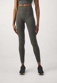 Tmavě zelené sportovní legíny s vysokým ribbed pasem. Hladká textura látky, přiléhavý střih, v kombinaci s černými sportovními botami.