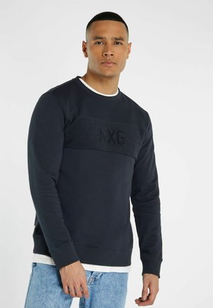 Marineblauwe trui met een gestructureerde horizontale band met "NXG" branding, ronde halslijn en geribbelde manchetten. Draagt over lichte spijkerbroeken.