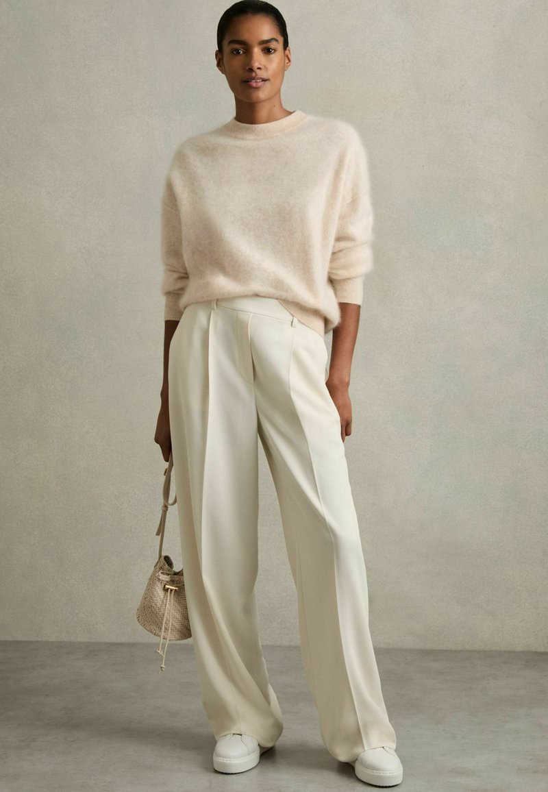 Helles beige Mohair-Pullover und weiße plissierte Hose. Das Model hält eine kleine geflochtene Tasche und trägt weiße Sneakers. Sanfte Texturen und neutrale Farben.