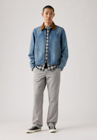 Τζιν μπουφάν με καφέ σουέντ γιακά, φορεμένο πάνω από καρό πουκάμισο, συνδυασμένο με ανοιχτό γκρι παντελόνια και μαύρα αθλητικά παπούτσια με λευκές σόλες.