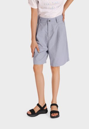 Persona che indossa pantaloncini azzurri chiari a vita alta, lunghezza al ginocchio, una t-shirt bianca con testo e sandali neri con punta aperta e suola spessa.