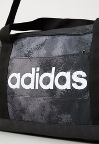 Čierna a šedá taška Adidas na dušu s veľkým bielym logom, čiernymi popruhmi a zipsom na vrchu.