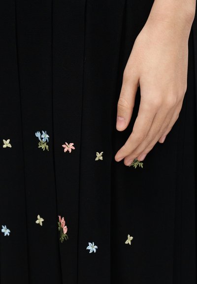 Jupe plissée noire avec broderie florale colorée en bleu, rose et vert. Une main touche le tissu, mettant en valeur la texture.