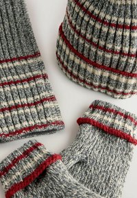 Strikket tilbehørssæt bestående af en grå hue og fingreløse handsker med røde og cremefarvede striber. Lavet af blødt uld, der fremhæver et struktureret mønster.