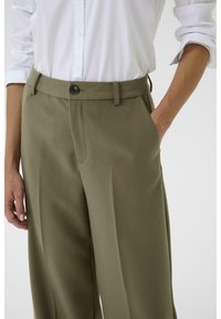 Khaki wijde broek met een gladde textuur, voorzien van een voorkeuro pocket, riemlussen en een knoopsluiting. Gecombineerd met een wit overhemd met knopen.
