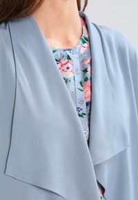 Blå, flödande ytterjacka med en drapera krage som bärs över en knappad blommig blus i ljusblått, med rosa och gröna blommönster.