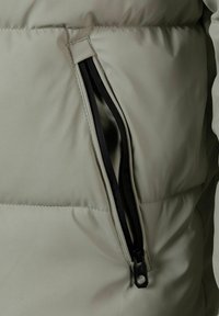 Chaqueta de abrigo de color gris claro con un bolsillo con cremallera visible. El bolsillo tiene cremalleras negras y una textura elegante y suave.