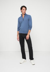 Blauwe trui met rits en lange mouwen, gemaakt van een zachte stof, gecombineerd met zwarte jeans en witte sneakers. Eenvoudig, modern ontwerp.
