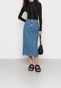 Svart långärmad topp med kontrasterande sömmar, parat med en medellång blå denimkjol och svarta sandaler. Håller en liten svart väska.