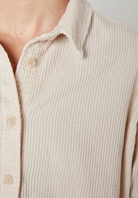 Light beige corduroy shirt with vertical ribbing, a buttoned placket, and a soft collar. Features rounded buttons and a textured surface.
