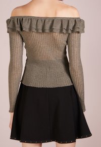 Schulterfreies Oberteil in olivgrünem Rippenstrick mit Rüschenausschnitt und langen Ärmeln. Kombiniert mit einem schwarzen, ausgestellten Rock mit goldenen Nieten.