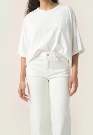Biała, oversize'owa koszulka z teksturowaną łatą z logo na piersi, w połączeniu z białymi dżinsami o wysokim stanie. Oba elementy w jednolitym kolorze.