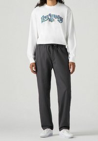 Bílá dlouhá košile s barevným logem "Levi Strauss & Co", kombinovaná s tmavě šedými elastickými kalhotami a bílými teniskami.