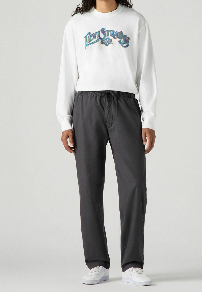 Bílá dlouhá košile s barevným logem "Levi Strauss & Co", kombinovaná s tmavě šedými elastickými kalhotami a bílými teniskami.