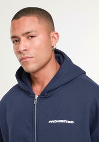 Navy zip-up hoodie with a hood, featuring a white "PROHIBITED" logo. Soft fabric with a smooth texture and silver zipper detail.