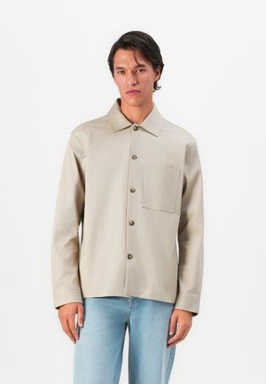 Man wearing beige buttoned jacket with chest pocket and light blue jeans, standing against plain white background.