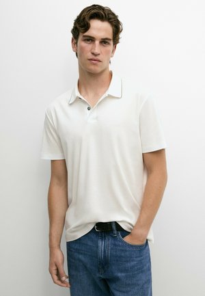 Giovane uomo con capelli castani corti indossa una polo bianca a maniche corte e jeans blu, in piedi con una mano in tasca su uno sfondo semplice.
