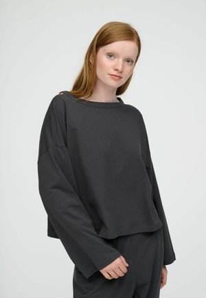 Noor naine punaste juustega, kandes lahtist tumedat halli pika varrukaga särki ja sobivaid pükse ühevärvilise halli tausta ees.