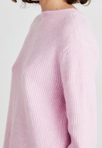 Osoba ubrana w miękki, jasnoróżowy sweter z prążkowanej dzianiny z długimi rękawami na tle gładkiej białej ściany.