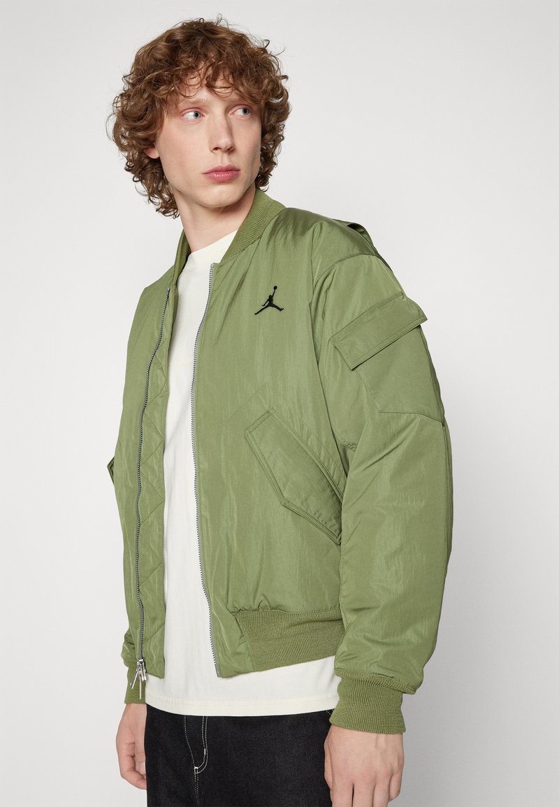 Chaqueta bomber en nylon verde oliva con cremallera frontal, puños de canalé y un logo negro en el pecho. Presenta bolsillos tipo parche en los lados.