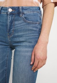 Hoch geschnittene blaue Jeans aus Denim mit glatter Textur, sanften Ausbleichungen, orangen Nähten und einer Fronttasche. Knopfdetail am Bund.