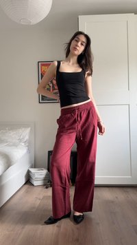 Νεαρή γυναίκα στέκεται σε ένα υπνοδωμάτιο φορώντας μαύρο αμάνικο τοπ, άνετο κόκκινο παντελόνι και μαύρα μυτερά παπούτσια, με ορατό ένα αφίσα και φωτιστικό.