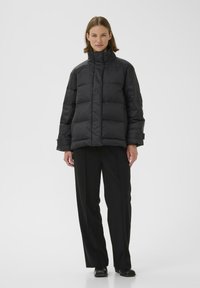 Sort pufferjakke med høj krave, lynlås lukning og quiltet design. Båret over sorte bukser, stoffet har en glat, skinnende tekstur.