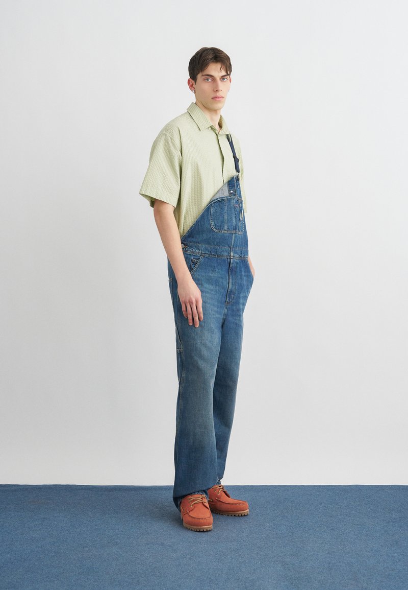 Junger Mann steht, trägt ein hellgrünes kurzärmeliges Hemd, blaue Jeanslatzhose mit einem gelösten Träger und braune Schuhe auf blauem Teppich.