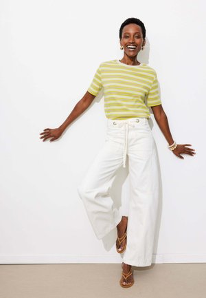 Femme souriante, appuyée contre un mur blanc, portant une chemise rayée jaune, un pantalon blanc ample, des sandales et des boucles d'oreilles créoles dorées.