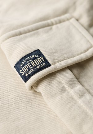 Tissu beige avec une texture côtelée et un ourlet plié, présentant une étiquette tissée bleue portant les inscriptions "SUPERDRY" et "TRADITIONAL WORK WEAR."