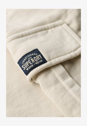 Tissu beige avec une texture côtelée et un ourlet plié, présentant une étiquette tissée bleue portant les inscriptions "SUPERDRY" et "TRADITIONAL WORK WEAR."