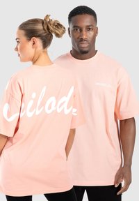 Lyserøde oversized t-shirts lavet af bomuld. Den ene har stort hvidt tekst på ryggen og et lille logo på forsiden. Rund halsudskæring.