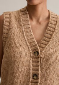 Beige knitted vest with a deep V-neck, ribbed neckline, and three dark buttons along the front. Textured wool material.