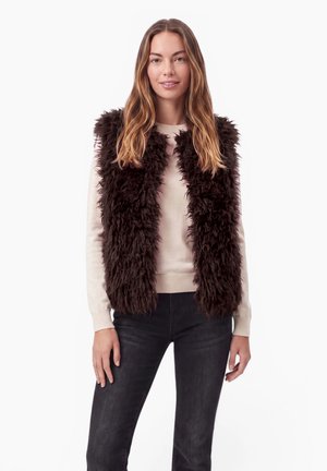 Femme aux longs cheveux bruns portant une veste en fourrure marron foncé sur un pull beige et un pantalon noir, debout devant un fond blanc.