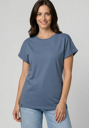 Frau mit schulterlangem dunklem Haar, die ein blaues T-Shirt mit kurzen Ärmeln und hellblaue Jeans trägt, lächelnd vor einem schlichten Hintergrund.