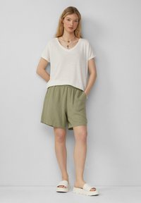 Biała koszulka z krótkim rękawem i dekoltem w serek zestawiona z oliwkowo-zielonymi szortami na gumce oraz kremowymi sandałami na koturnie. Prosty design, swobodne dopasowanie.