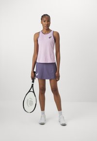 Tennisantrekk med en lysebrun ermeløs topp med et geometrisk mønster, kombinert med en lilla skjørt og holder en racket.
