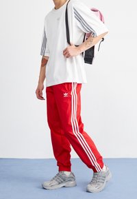 Röda sportbyxor med vita ränder, vit oversize-t-shirt med grå ränder, grå sneakers och en rosa och svart ryggsäck.