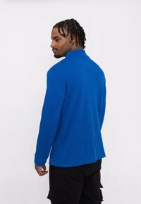 Pull bleu texturé avec motif en relief, col montant et manches longues. Porté avec un pantalon cargo noir, mettant en valeur une coupe décontractée.