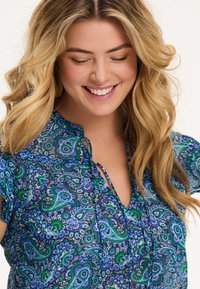 Smaidoša sieviete ar viļņainiem gaišiem matiem, valkājot zilu, zaļu un purpura ziedu rakstu blūzi ar atslēgas caurumu dekoltē.