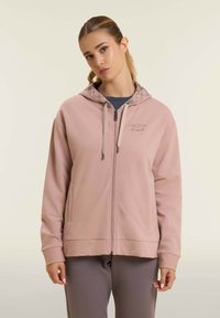 Felpa con zip di colore rosa chiaro realizzata in tessuto morbido, con fodera del cappuccio a fantasia, due tasche frontali e dettagli con cordoncino.