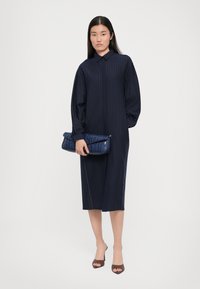 Marineblå pinstriped kjole med krage, lange ærmer og mellemlangt forkant. Modellen holder en tekstureret blå clutch og har brune åbne sko på.