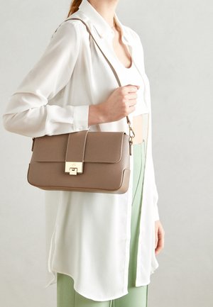 Donna che indossa una camicia bianca lunga e pantaloni verde chiaro tiene una borsa a tracolla color taupe con fibbia dorata sulla spalla.