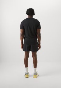 Camiseta negra de manga corta, pantalones cortos negros texturizados y zapatillas gris-amarillas. Calcetines blancos hasta el tobillo. Vista desde la parte de atrás contra un fondo liso.