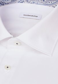 Col de chemise blanche et boutons avec une étiquette de marque "seidensticker" visible sur un fond bleu et blanc à motifs.