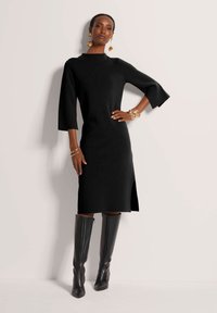 Schwarzes gestricktes Kleid mit hohem Kragen, dreiviertellangen Ärmeln und seitlichem Schlitz. Kombination mit knielangen schwarzen Lederstiefeln.