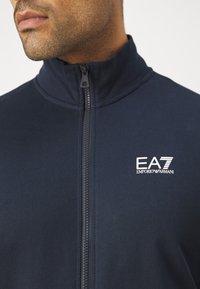 Giacca a zip blu navy realizzata in tessuto morbido, con colletto alto e zip frontale. Include il logo EA7 Emporio Armani in bianco sul petto.